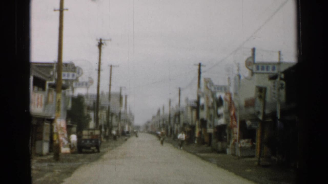 1952 Japan home-movie frame looking along a town street lined with shopfronts, utility poles, hanging signs, and a small truck at the curb.