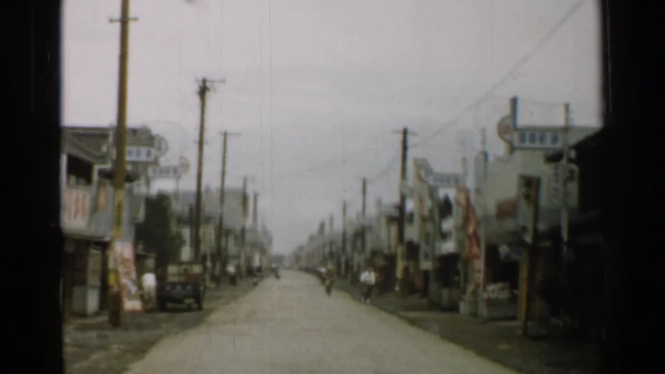 1952 Japan home-movie frame looking along a town street lined with shopfronts, utility poles, hanging signs, and a small truck at the curb.