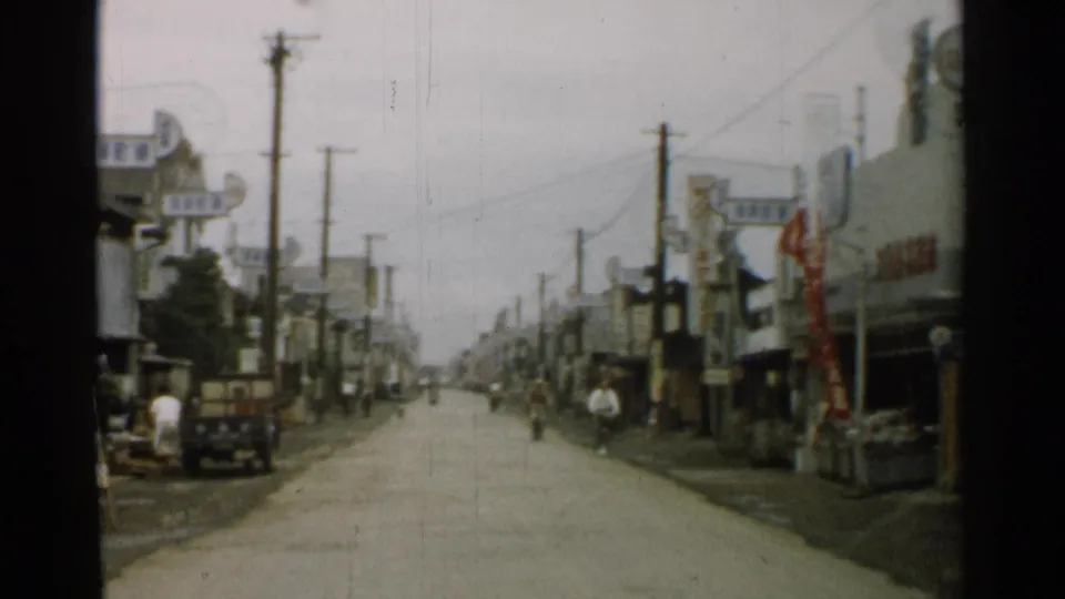 1952 Japan street view from a moving vehicle, with storefront signs, utility poles, a small truck, and pedestrians along the roadside.