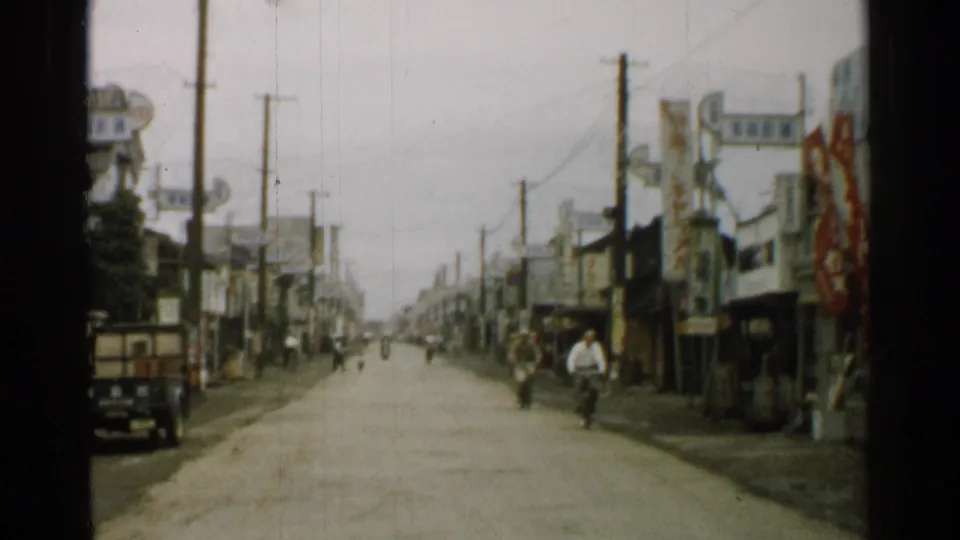1952 Japan home-movie frame showing a closer town street view with shopfronts, a small truck, pedestrians, signs, poles, and roadside banners.