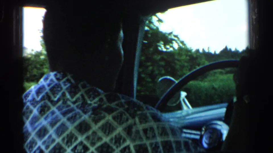 1952 Japan driving-view frame from inside a vehicle, showing a man in a patterned shirt at the wheel with trees and roadside greenery outside.