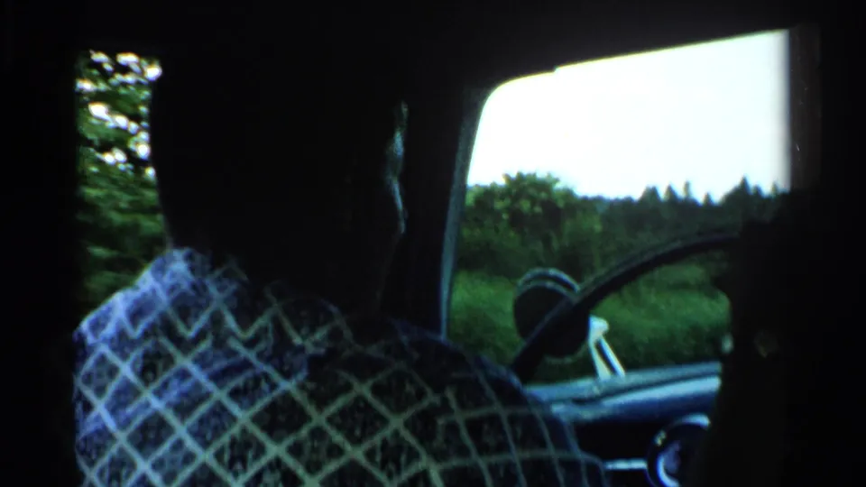 Backseat view of a silhouetted male driver in 1952 Japan, with a large steering wheel and green countryside visible through the side window.