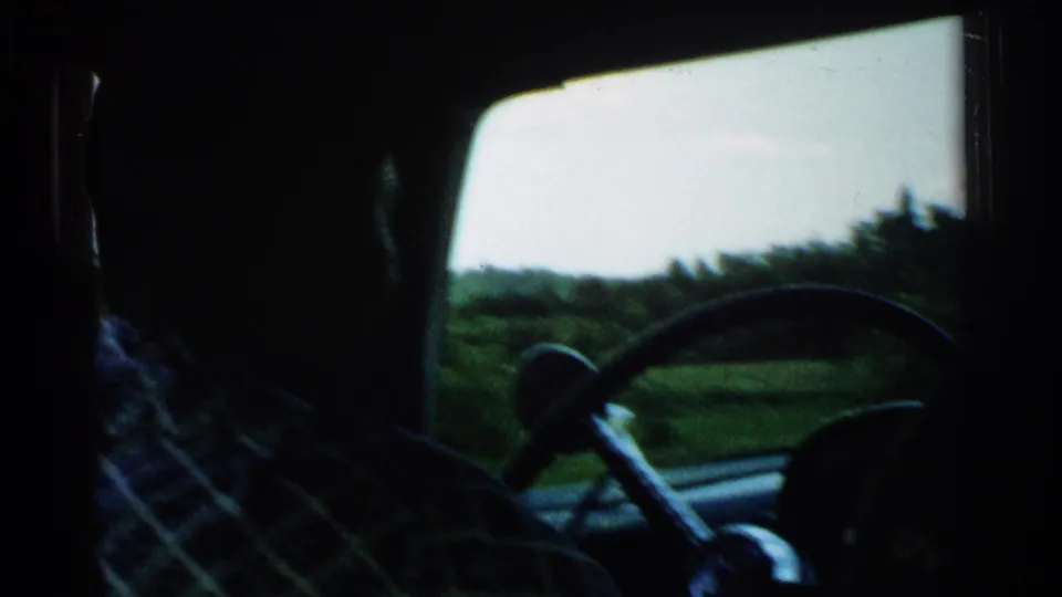 The driver and steering wheel are seen from the rear seat, with a soft rural landscape outside the window in 1952 Japan footage.