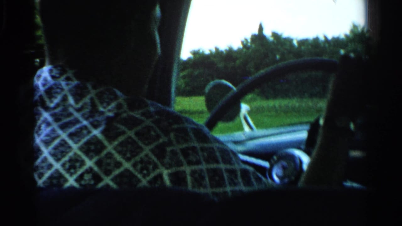 Driver seen from behind steering a car past green roadside fields.