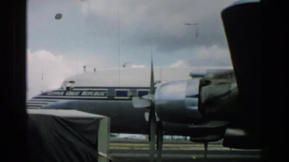 The parked propeller aircraft remains in view on the tarmac, partly framed by dark foreground objects and film-edge shadow.
