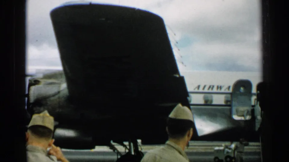 At a Japan airfield in 1952, uniformed men stand near a dark aircraft structure while a white plane with an open door appears in the background.