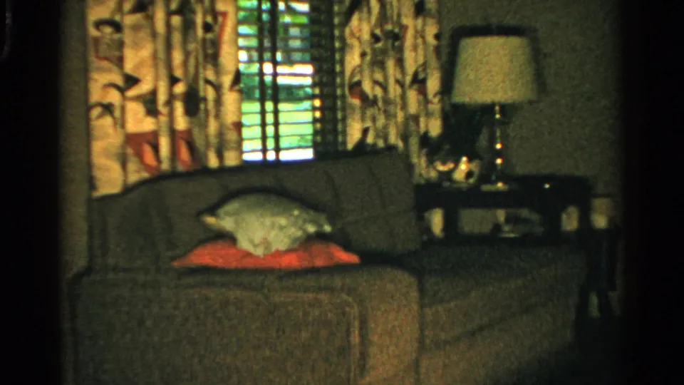 1950s domestic interior with a couch, pillows, patterned curtains, shuttered window, side table, and shaded lamp.