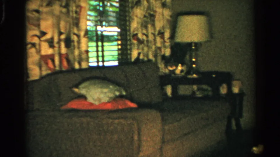 1952 Japan home-movie frame showing a couch with red and pale pillows near drapes, blinds, and a lit side table lamp.