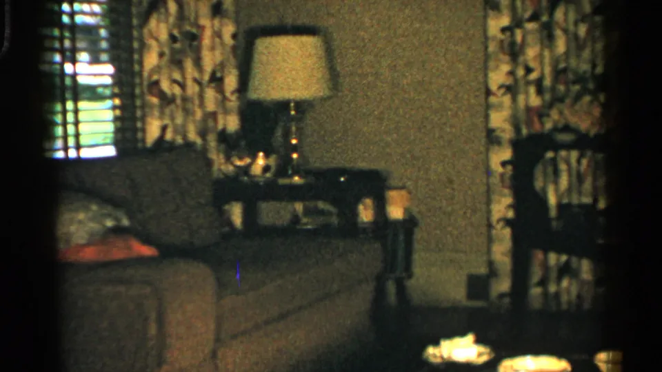 1952 Japan living room frame centered on a lamp and side table beside a couch, with drapes, blinds, and small foreground objects.