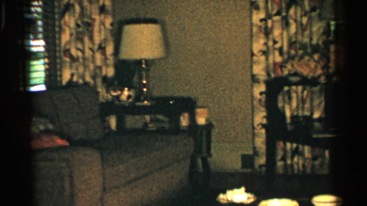 1952 Japan home-movie frame of a dim living room with sofa, patterned curtains, lamp, side table, and small objects on a foreground table.