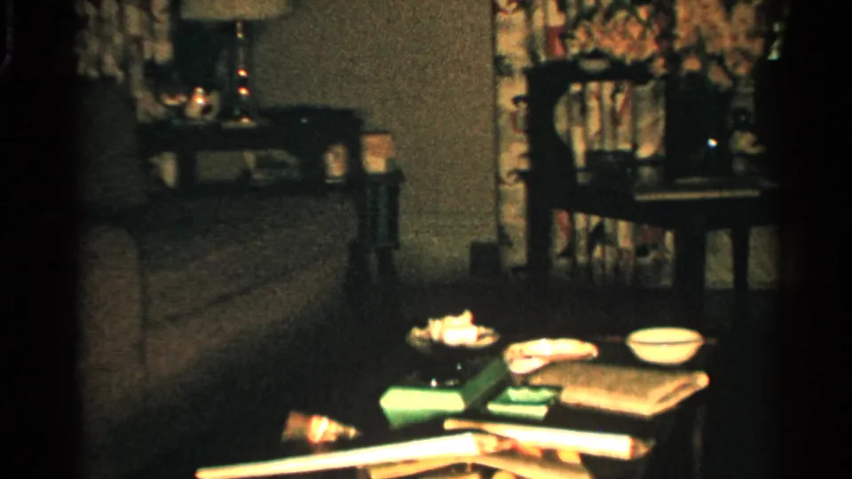 1952 Japan home-movie still showing a sofa, wooden chair, patterned curtains, and a foreground table holding bowls, papers, and small items.