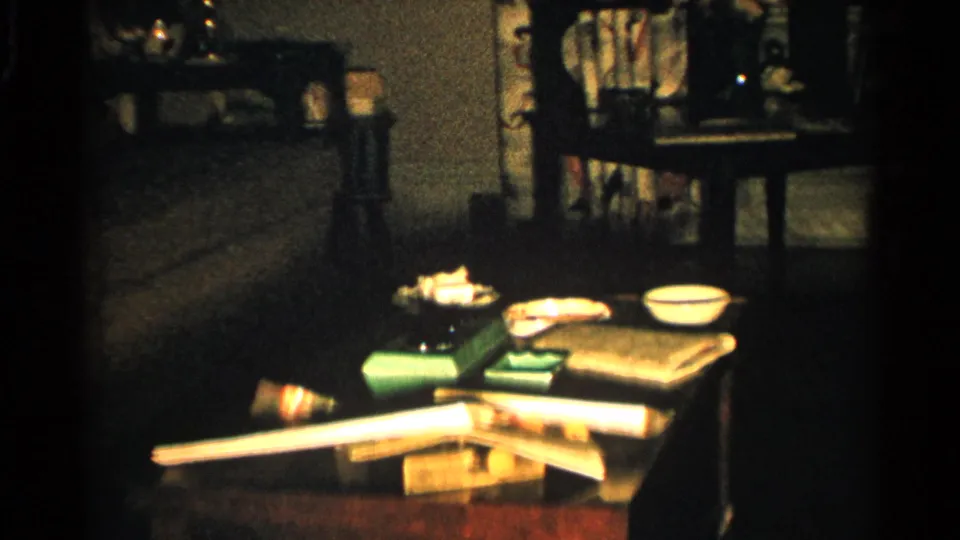 1952 Japan interior frame with a cluttered low table, bowls, books or papers, long pale object, sofa, chair, and patterned curtains.