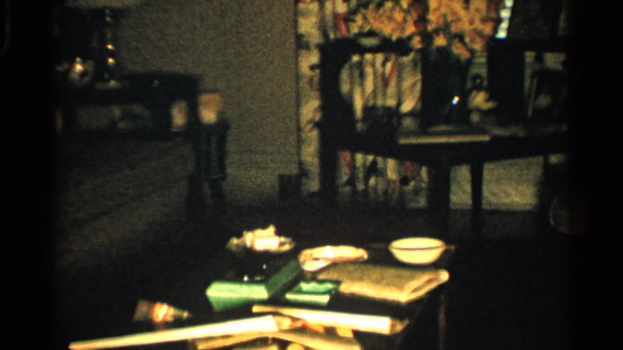 Dim living room with a low table, bowls, books, curtains, chairs, and framed photos.