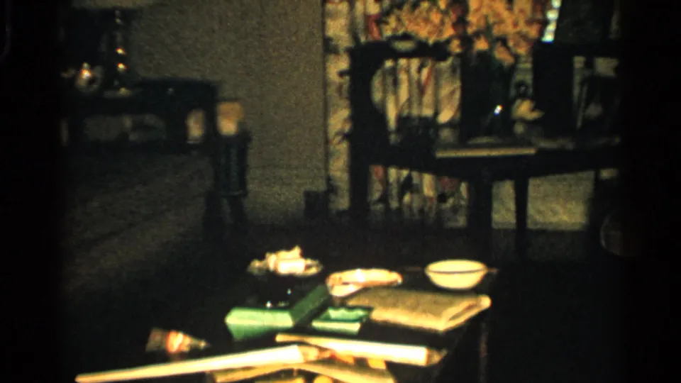 1952 Japan home-movie frame showing a dim family living room with a low table, household objects, floral curtains, chairs, and photographs.