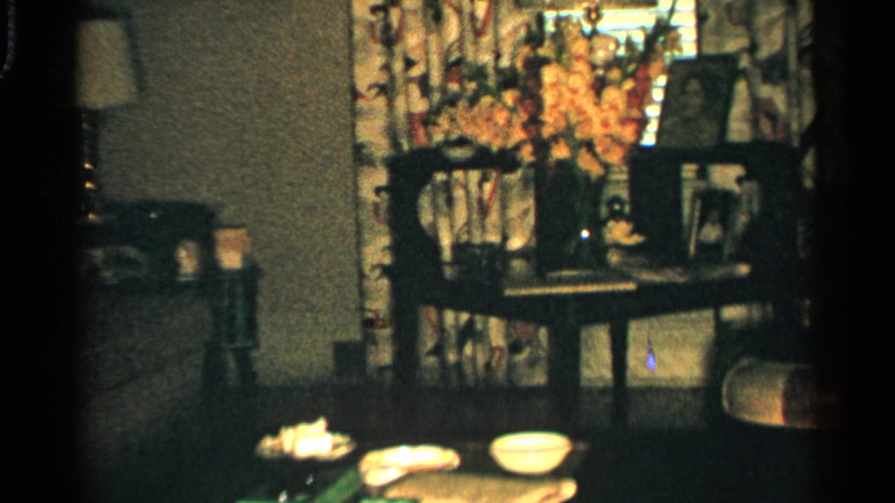 Living room corner with a table, bowls, floral curtains, orange flowers, lamp, and framed photos.