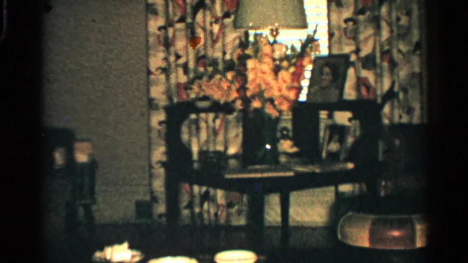 1952 Japan home-movie still showing a side table with flowers, lamp, framed photographs, floral curtains, and nearby seating in a private living room.