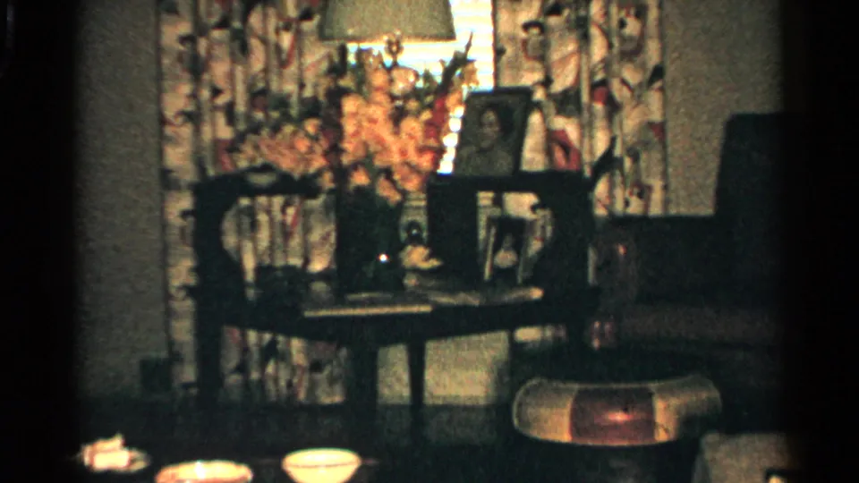 1952 Japan domestic interior with flowers, framed photographs, a lamp, floral curtains, table objects, and chairs in soft 8mm light.