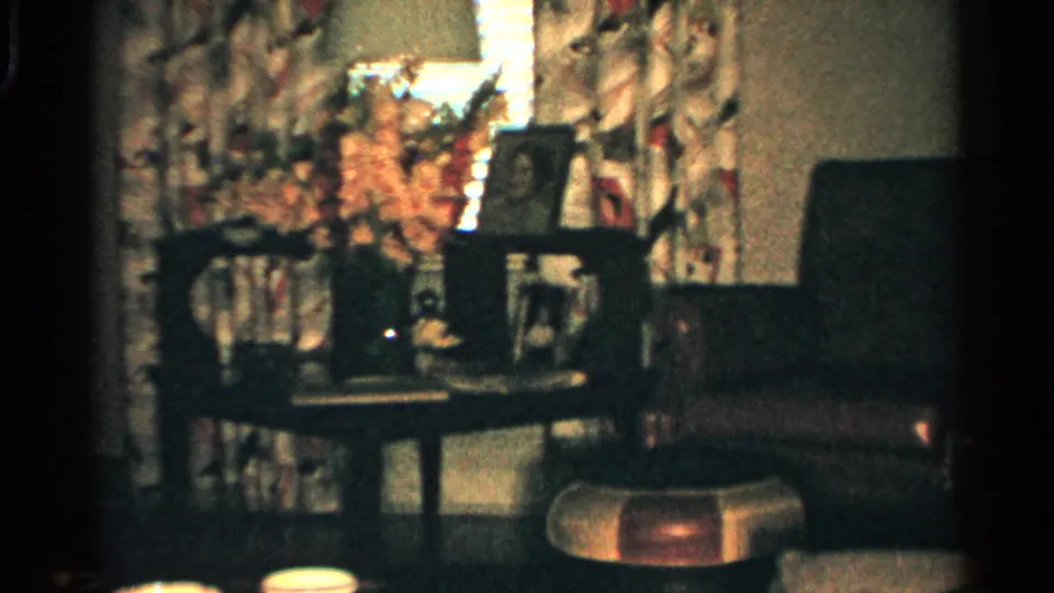 1952 Japan home-movie frame showing a softly lit living room with floral curtains, framed photos, flowers, chairs, and a low table.