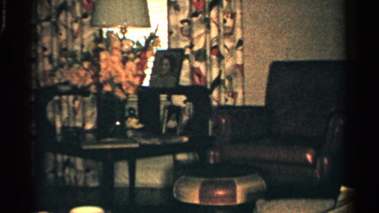 Living room corner with lamp, curtains, framed photos, side table, flowers, and chairs.