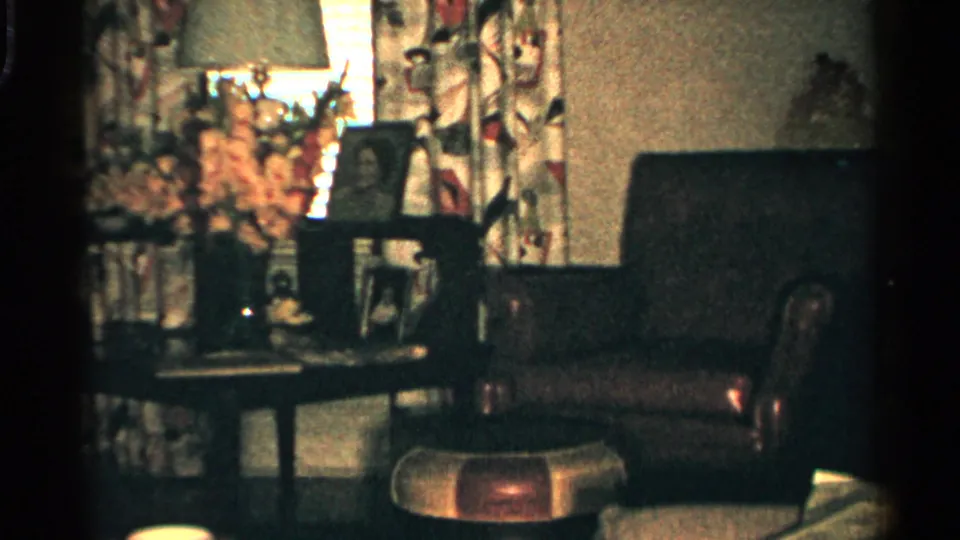 1952 Japan home-movie view of a private living room with floral curtains, table flowers, framed photos, seating, and a low table.