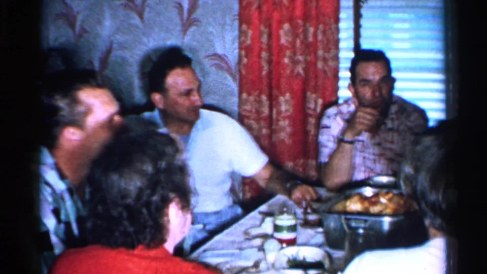 1952 Japan home-movie frame of adults gathered at a dining table with food, glasses, curtains, and window blinds visible.