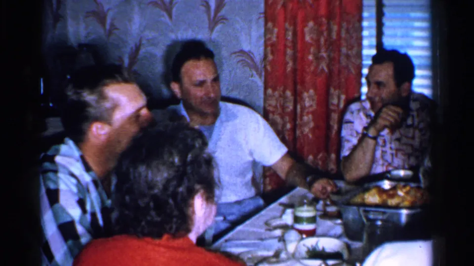 In a 1952 Japan home-movie frame, adults sit around a dining table with a roasted dish, cups, red curtains, and patterned wallpaper.