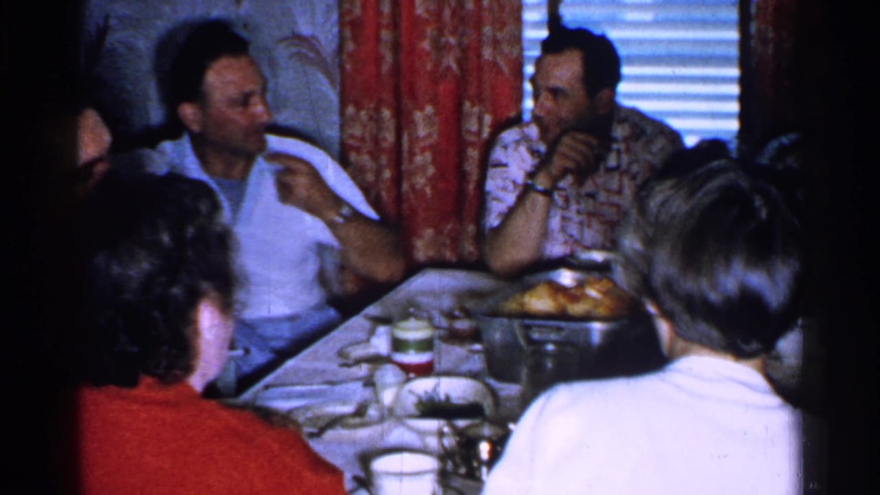 Several adults sit at a table while a man gestures and another listens near a dish of food.