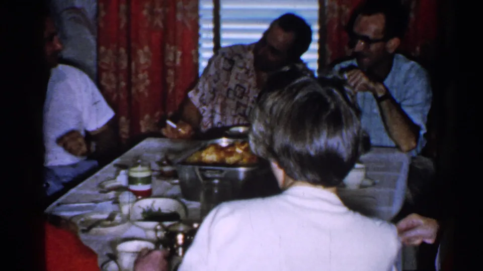 The handheld frame shows adults seated at a home dinner table, with food in a pan, cups, red curtains, and a foreground figure from behind.