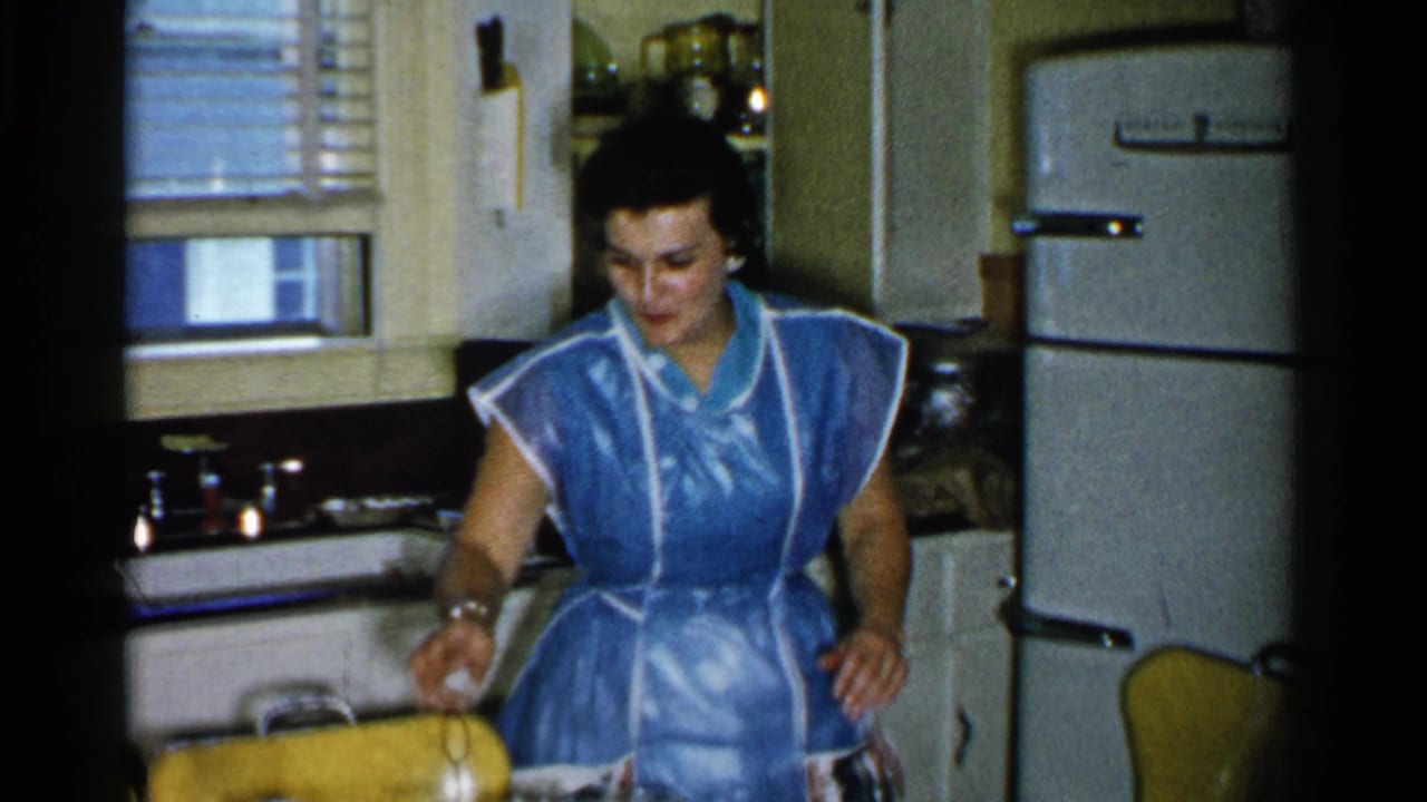 In 1952 Japan home-movie footage, a woman in a blue apron works near the sink in a small kitchen with a refrigerator and yellow chairs.