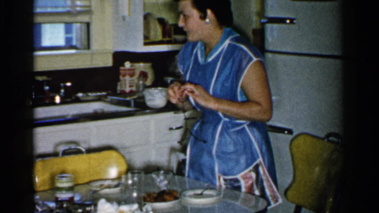Woman in a blue apron stands by a kitchen table with dishes, glassware, and a refrigerator behind her.