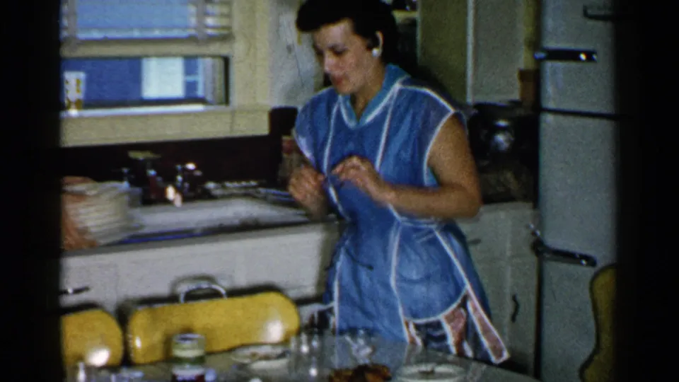 In a private kitchen, the woman in a blue apron handles a small item near dishes, a sink counter, window blinds, and a refrigerator.