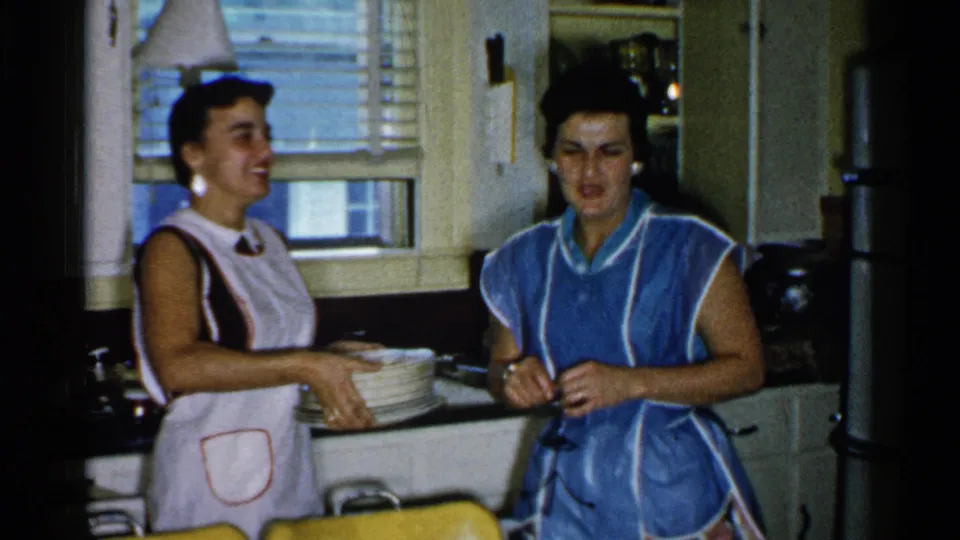 Two women in aprons share a brief kitchen moment in 1952 Japan home-movie footage, with dishes, a window, and cabinets visible.