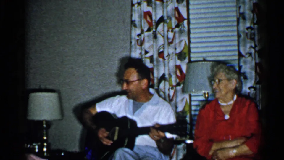 1952 New Hampshire home-movie frame of a man holding a guitar beside an older woman in a living-room setting.