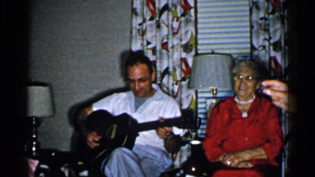 Man seated with an acoustic guitar beside an older woman in a living room.