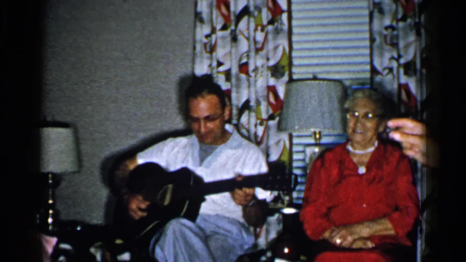 1952 New Hampshire home-movie frame of a man playing guitar while an older woman in red sits beside him indoors.