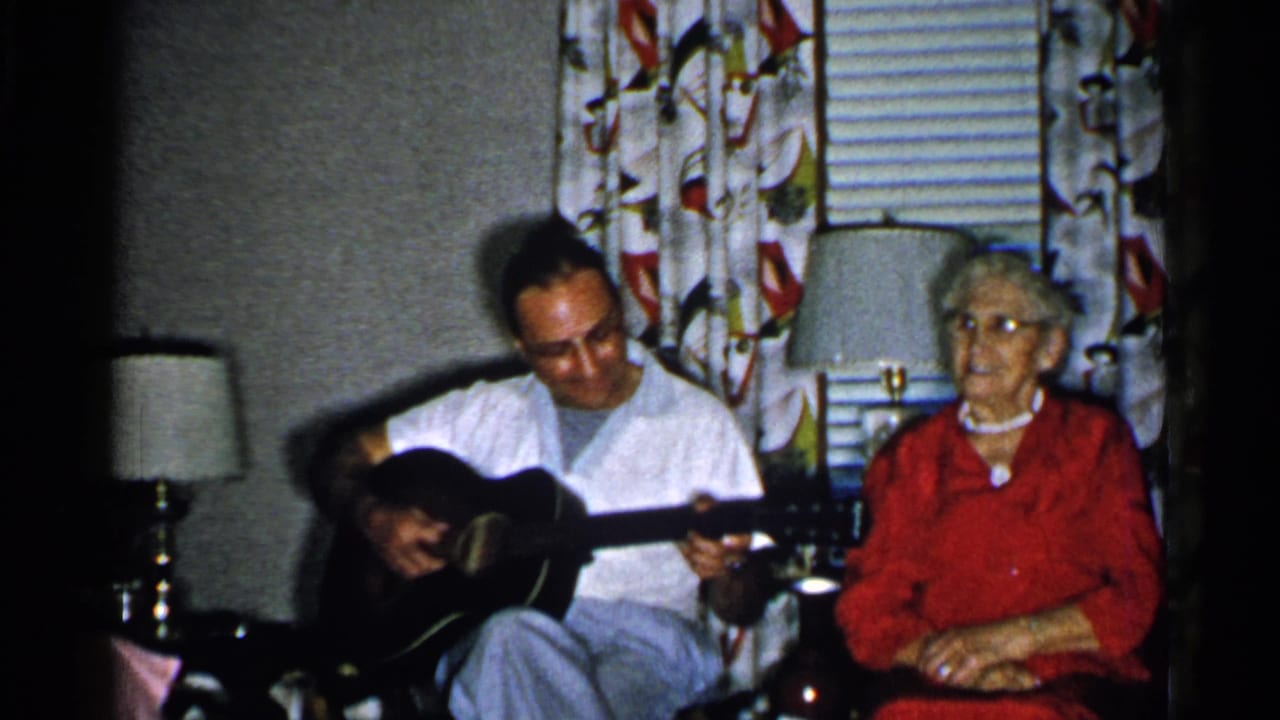 Man looks down at an acoustic guitar while an older woman sits beside him.