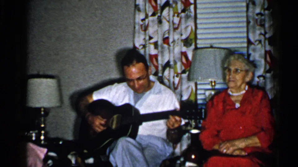 Home-movie frame showing a seated guitarist and older woman in a 1952 New Hampshire living room with lamps and drapes.