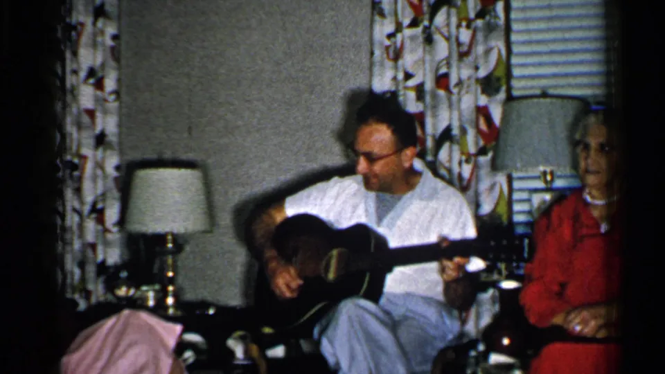 New Hampshire 1952 home-movie frame with a man holding guitar and an older woman seated near the edge of a living room scene.