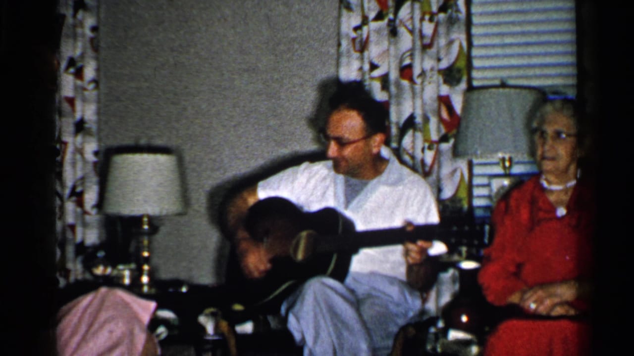 1952 New Hampshire home-movie frame showing a man playing acoustic guitar beside an elderly woman in red in a private living room.