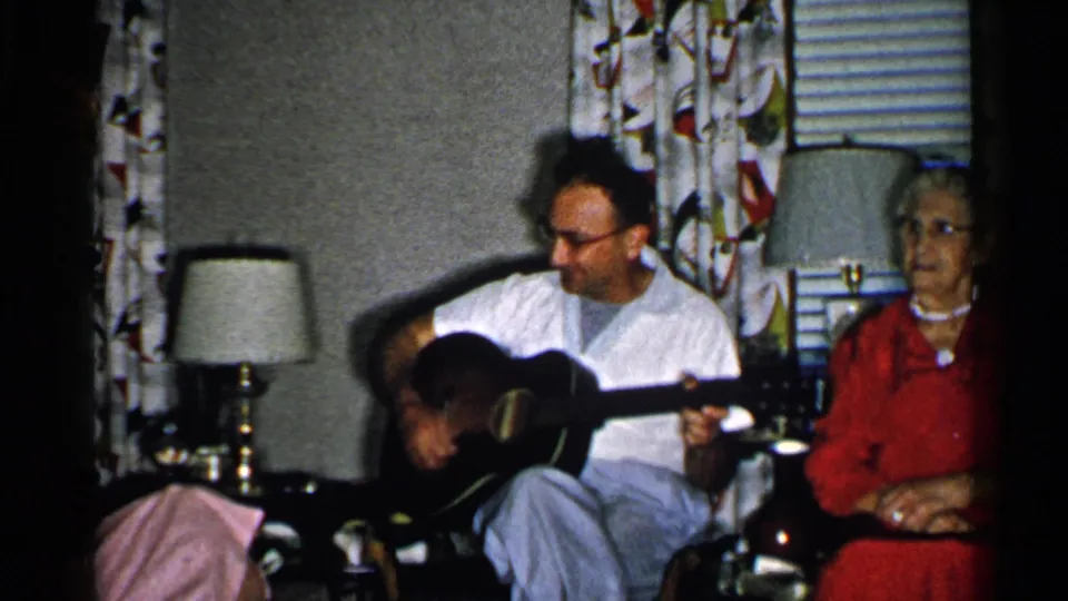 1952 New Hampshire home-movie frame showing a man playing acoustic guitar beside an elderly woman in red in a private living room.