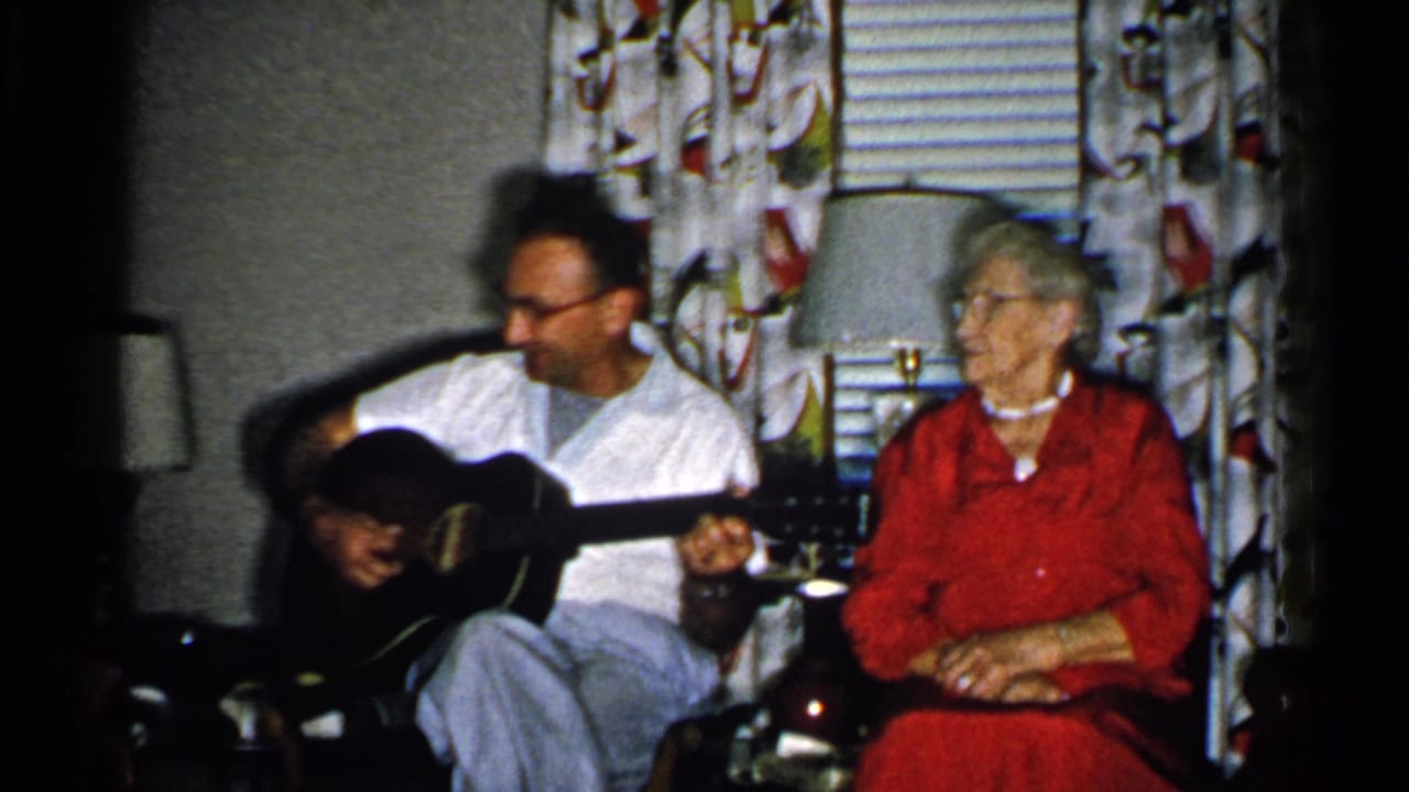 Man plays guitar while an elderly woman turns toward him in a living room.