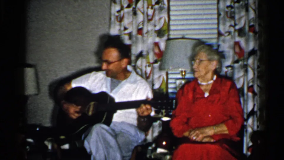 1952 New Hampshire frame with a seated guitarist and elderly woman in red near patterned curtains, blinds, and lamps.