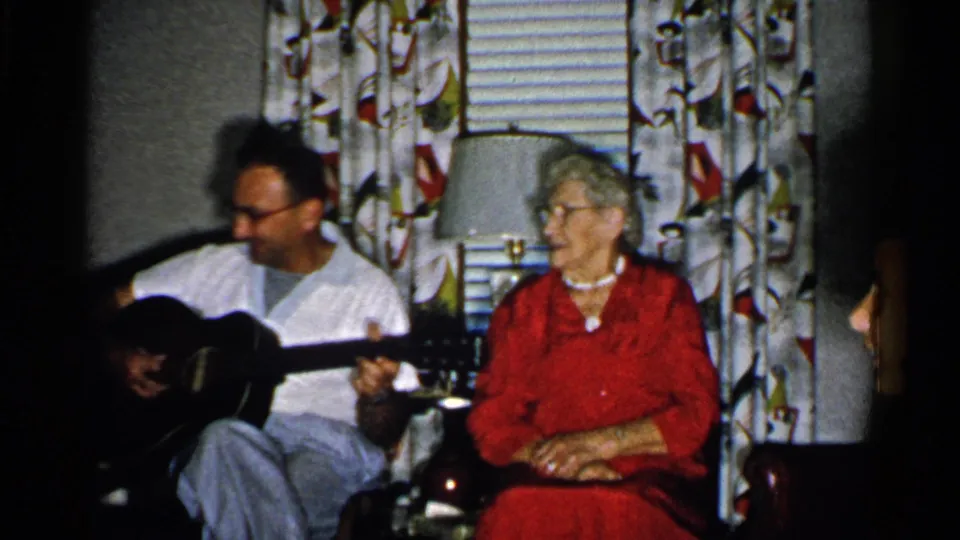1952 New Hampshire home-movie frame showing a man with an acoustic guitar next to an elderly woman in a mid-century living room.