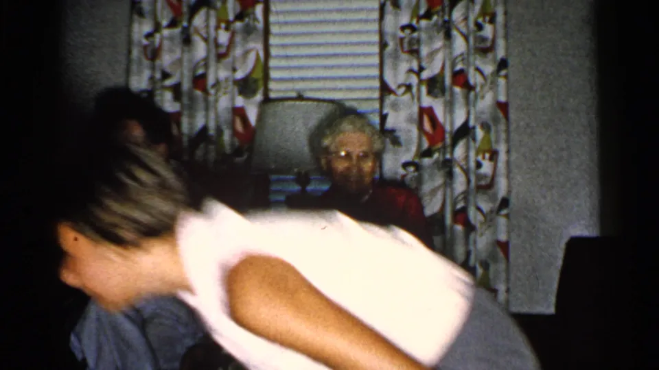 1952 New Hampshire home-movie frame with a child crossing the foreground before a seated guitarist and elderly woman in a living room.