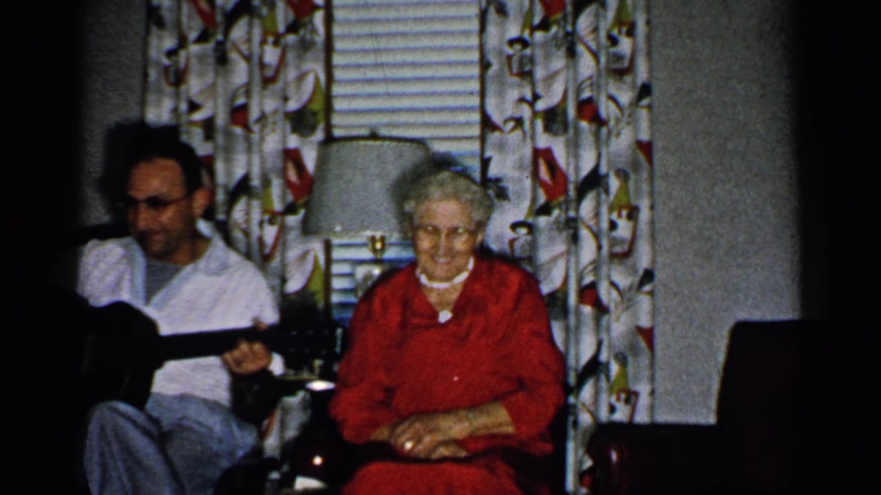 1952 New Hampshire home-movie frame showing a man with an acoustic guitar seated beside an older woman in red near curtains and a lamp.