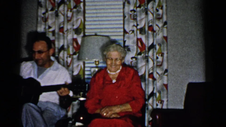 1952 New Hampshire home-movie frame showing a man with an acoustic guitar seated beside an older woman in red near curtains and a lamp.