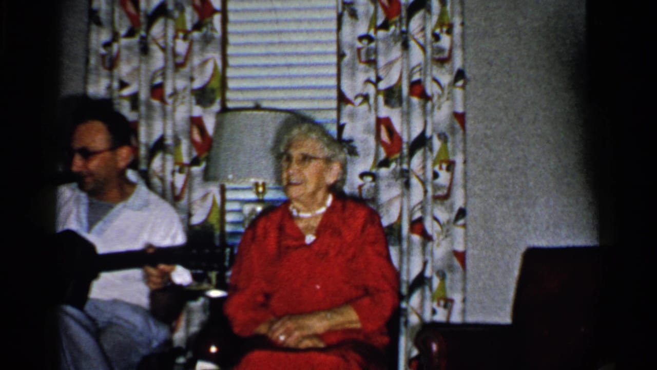 Older woman in red turns toward a man holding an acoustic guitar indoors.