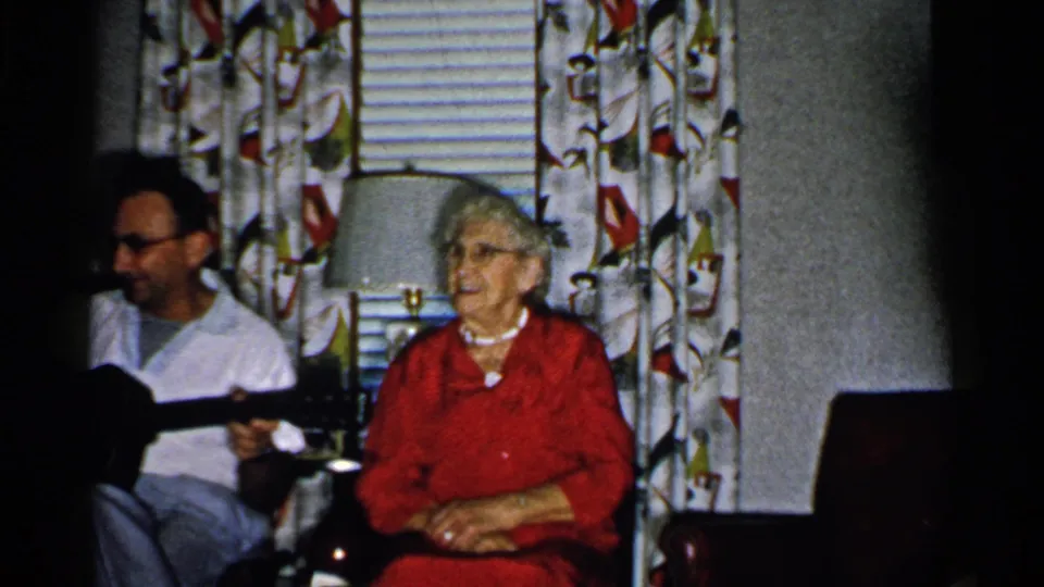 1952 New Hampshire frame with an older woman in red looking toward a seated guitar player in a mid-century living room.