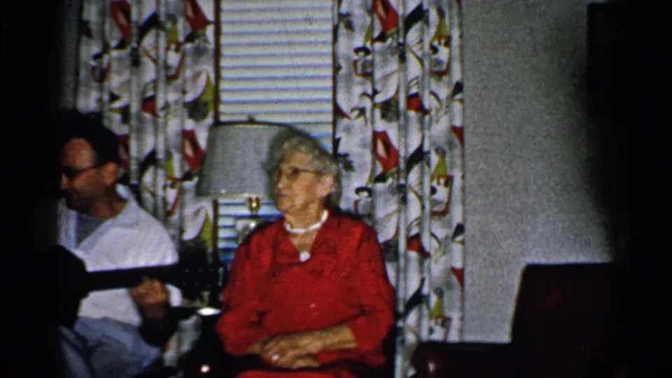 1952 New Hampshire home-movie frame showing a man holding a guitar and an older woman seated close by in a private interior.