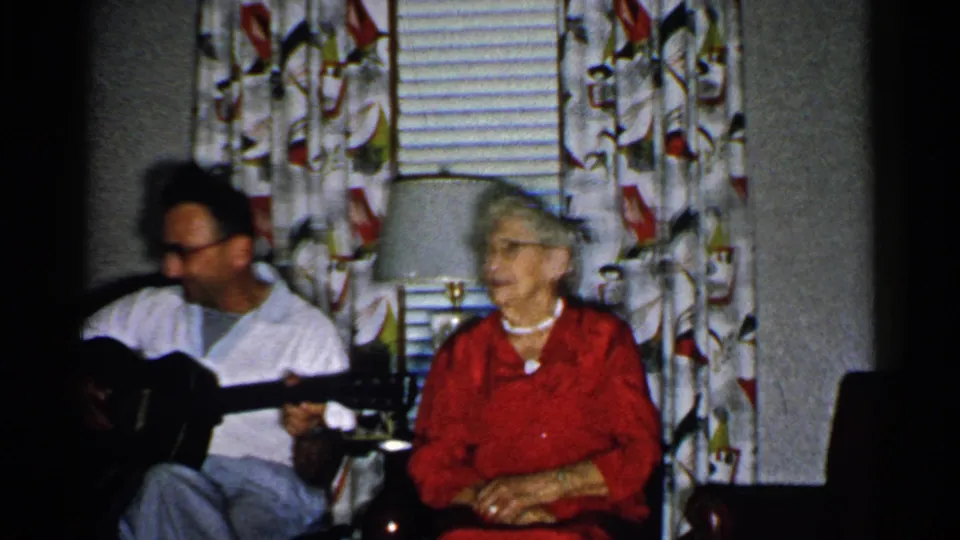 1952 New Hampshire home-movie frame of a seated older woman listening beside a man with an acoustic guitar in a dim living room.